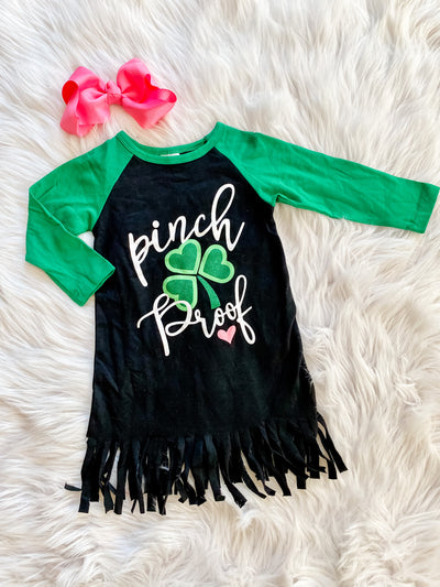 Girls long sleeve tshirt style dress with green sleeves, black dress with fringe hem, and the words Pinch Proof on the front with a shamrock and heart. 
