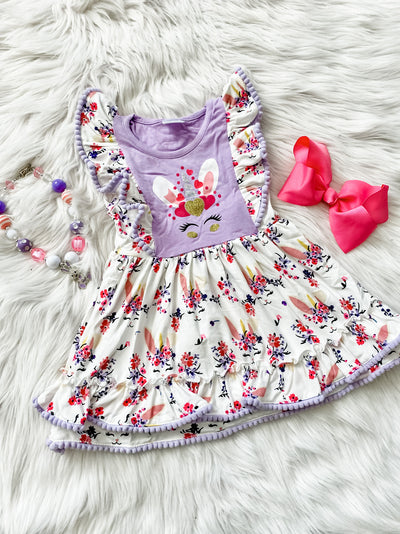 Girls flutter sleeve dress with floral unicorn bunny pattern and purple pom poms along the hem. 