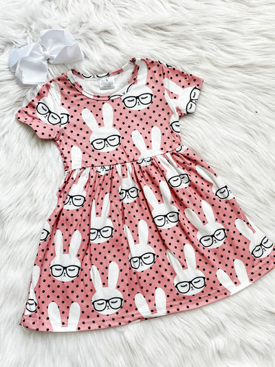 Blush colored short sleeve girls dress with black polka dots covered in white bunnies wearing black glasses.