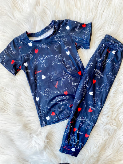 Unisex kids two piece pajamas with short sleeve and pants that are navy blue with white outlined dinosaurs and red and white hearts. 